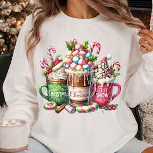 May include: A white long-sleeve sweatshirt featuring a graphic print of three mugs filled with hot cocoa, whipped cream, candy canes, and colorful marshmallows. The mugs are decorated with the text "CHRISTMAS", "Christmas", and "LET IT SNOW".