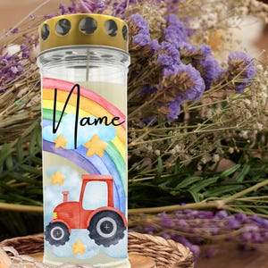May include: A cylindrical candle with a gold-colored lid. The candle features a watercolor design of a red tractor, rainbow, clouds, and stars. The word "Name" is written in cursive. The candle is surrounded by purple flowers.