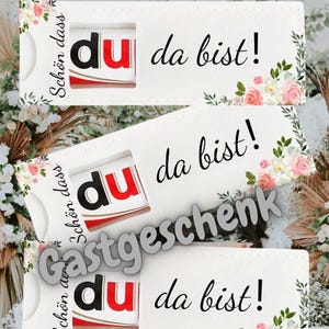 May include: White gift card holders with floral accents and the text "du da bist!" in black. The holders feature a cutout revealing a red and white product. The word "Gastgeschenk" is also visible.