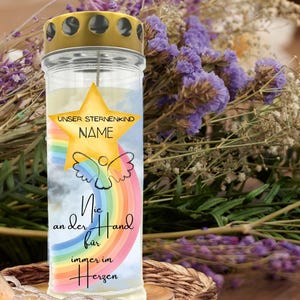 May include: A glass candle with a gold-colored lid. The candle has a star and rainbow design with the words "UNSER STERNENKIND NAME" and other text. The candle is surrounded by purple flowers.