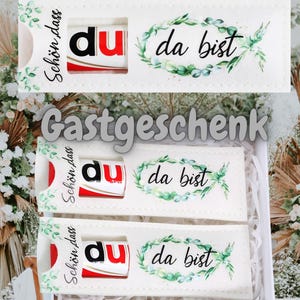 May include: A gift box containing three white rectangular packages, each holding a chocolate bar. The packages feature the text "Schön, dass du da bist" and a green floral design. The word "Gastgeschenk" is also visible.
