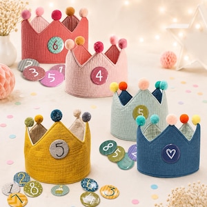 May include: A collection of fabric crowns in red, pink, light blue, mustard yellow, and dark blue, each topped with small pom-poms. The crowns feature circular badges with numbers or a heart symbol, accompanied by loose circular number badges.