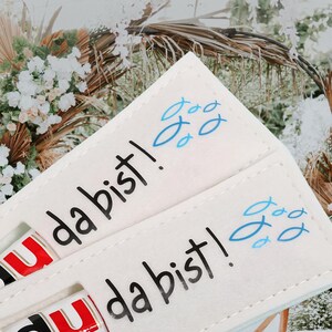 May include: Cream-colored felt holder with the text "da bist!" in black and blue fish illustrations. The holder contains a Nutella stick. The background features a blurred floral arrangement.