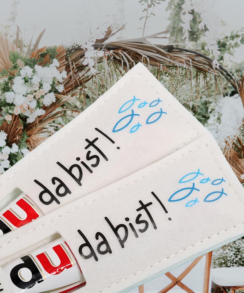 May include: Cream-colored felt holder with the text "da bist!" in black and blue fish illustrations. The holder contains a Nutella stick. The background features a blurred floral arrangement.