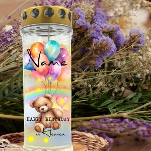 May include: A glass candle holder with a gold-colored lid. The candle features a design with colorful balloons, a rainbow, and a teddy bear. Text on the candle reads "Name", "Happy Birthday", and "in Heaven".
