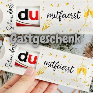 May include: A white gift tag with the words "Schön, dass du mitfeierst" and "Gastgeschenk" in black and red. The tag features a small window with the word "du" and celebratory graphics.