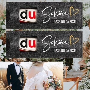 May include: Two dark gray gift boxes with the word "du" in a red and white design. The boxes have the text "Sch&ouml;n, DASS DU DA BIST!" in white script with a gold heart. The image also shows a bride holding a bouquet and a groom in a black suit.