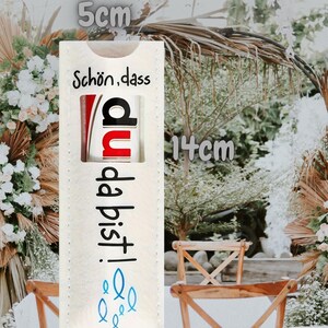 May include: A rectangular, off-white fabric item, 5 cm wide and 14 cm tall, with German text and a graphic. The text reads "Sch&ouml;n, dass du da bist!" in black, with a red and black graphic. Blue fish icons are at the bottom.