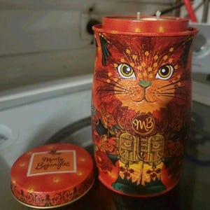 May include: A cylindrical orange metal tin featuring a stylized cat illustration with large yellow eyes and a gold MB logo. The tin has a matching lid with the text Monty Bojangles printed in a white square. The container holds a candle with two wicks.