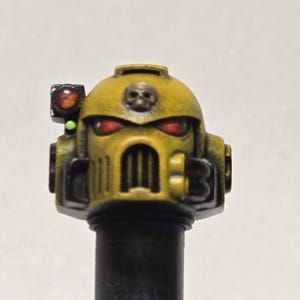 May include: Close-up of a yellow and black helmet with a skull emblem and red eye lenses. The helmet has a detailed design with a vertical grill and a small light on the side. The helmet is mounted on a black cylindrical base.