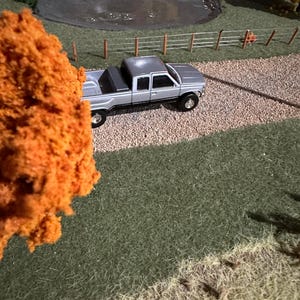 May include: A silver pickup truck with a black bed is parked on a gravel road. The truck is surrounded by green grass and a small pond. There is a fence in the background.
