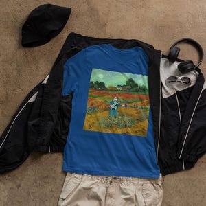 Op de afbeelding: Een blauw T-shirt met een schilderij van een persoon die gitaar speelt in een bloemenveld. Het schilderij bevindt zich in een rechthoekige lijst en is gecentreerd op het shirt. Het shirt ligt op een zwart-wit jack met een zonnebril en koptelefoon.