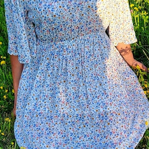 May include: A light blue floral print dress with a gathered waist and elbow-length sleeves. The dress is made of a lightweight fabric and is worn with a straw hat. The dress is surrounded by green grass and yellow flowers.