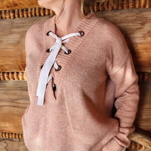 May include: A light pink sweater with white laces tied in a criss-cross pattern at the neckline. The sweater is made of a soft, fuzzy material and has long sleeves.
