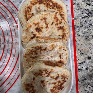 May include: Four pieces of flatbread arranged on a white oval plate. The bread has a light tan color with darker brown spots, suggesting it's been baked. The plate sits on a red and white patterned surface.