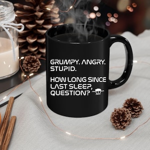 May include: Black ceramic mug with white text: "GRUMPY. ANGRY. STUPID. HOW LONG SINCE LAST SLEEP, QUESTION?" The mug has a curved handle and is likely used for hot beverages. Steam rises from the mug, suggesting a warm drink.