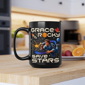 Puede incluir: Taza de cerámica negra con el texto "GRACE ROCKY SAVE STARS" y un gráfico espacial. El diseño incluye un astronauta, planetas y un cohete. La taza tiene un asa grande.