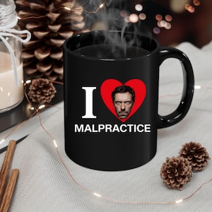 Puede incluir: Una taza de cerámica blanca con un asa negra y el texto "But First, Coffee" en negrita negra. La taza se encuentra sobre un fondo borroso, sugiriendo un ambiente acogedor.