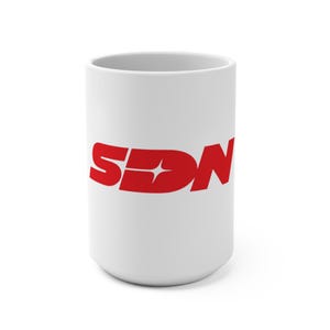 Könnte beinhalten: Eine weiße Keramiktasse mit einem roten "SDN"-Logo. Das Logo enthält ein Stern-Design in den Buchstaben. Die Tasse ist zylindrisch und für Heißgetränke geeignet. Das Design ist schlicht und auffällig.