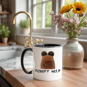 Può includere: Una tazza in ceramica bianca con manico e bordo neri. La tazza presenta un volto di cartone animato con grandi orecchie e bocca, con le parole "ROBERT HELP" stampate sotto. La tazza è su una superficie di legno.