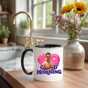 May include: White ceramic mug with a black handle and rim. The mug features a graphic of a man with two pink heart-shaped balloons and the words "GOOD MORNING" in blue. The mug is on a wooden surface.