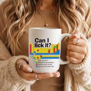 May include: A white ceramic mug featuring a periodic table graphic with the text Can I lick it? and a colour-coded legend. The legend indicates yellow for sure, go for it, green for maybe, not the best idea, red for please, don't do that, and blue for see you in the other side.