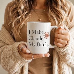 May include: A white ceramic mug featuring the dark gray text "I Make Claude My Bitch." printed on the front, accompanied by a small, stylized terracotta-colored sunburst graphic positioned to the right of the word "Claude."
