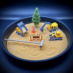 May include: A miniature construction site scene in a black tray. The scene includes a yellow dump truck, excavator, and bulldozer, a small green tree, orange traffic cone, and a construction barrier. The sand is raked with circular patterns.