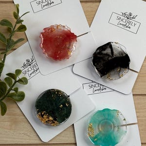 May include: Four resin phone grips in red, black, green, and teal, each with gold leaf accents. The grips are on white cards with the text "Sincerely LoveAsher". The grips have a unique, artistic design.
