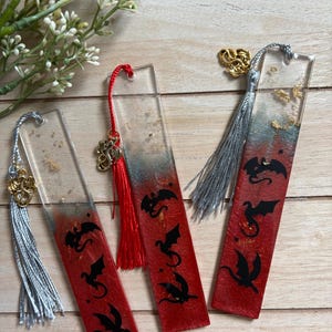 May include: Three rectangular resin bookmarks featuring a red and grey gradient base with embedded gold flakes. Each bookmark displays black dragon silhouettes and includes a decorative tassel with a small gold-toned dragon charm attached to the top.