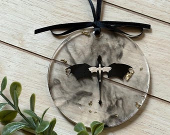 May include: A circular, transparent ornament featuring a black dragon silhouette and gold leaf details. It has a black ribbon for hanging. The background has a smoky grey design.