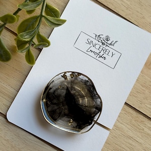Puede incluir: Un soporte para tel&eacute;fono de resina transparente con un dise&ntilde;o negro y dorado, sobre una tarjeta blanca con el texto "Sincerely LoveAsher". El agarre tiene un borde dorado y est&aacute; colocado sobre una superficie de madera con una planta verde en el fondo.