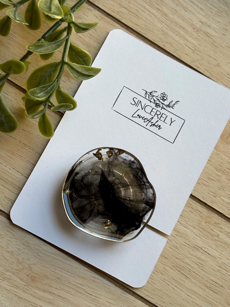 Puede incluir: Un soporte para tel&eacute;fono de resina transparente con un dise&ntilde;o negro y dorado, sobre una tarjeta blanca con el texto "Sincerely LoveAsher". El agarre tiene un borde dorado y est&aacute; colocado sobre una superficie de madera con una planta verde en el fondo.