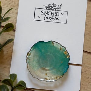 Puede incluir: Un soporte para tel&eacute;fono de resina turquesa y dorada con borde transparente, sobre una tarjeta blanca. La tarjeta tiene el texto "Sincerely LoveAsher" en negro. El soporte para tel&eacute;fono tiene detalles de pan de oro.