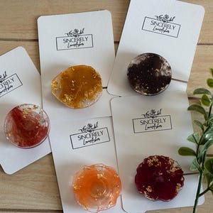 May include: A collection of colorful resin phone grips with gold leaf accents, displayed on white cards with the text "Sincerely Love Asher". The grips are in shades of red, orange, brown, and yellow.