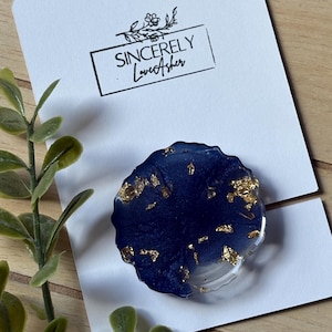Puede incluir: Un soporte para tel&eacute;fono de resina azul marino con detalles de pan de oro. El agarre redondo tiene una base transparente y est&aacute; sobre una tarjeta blanca con el texto "Sincerely LoveAsher".