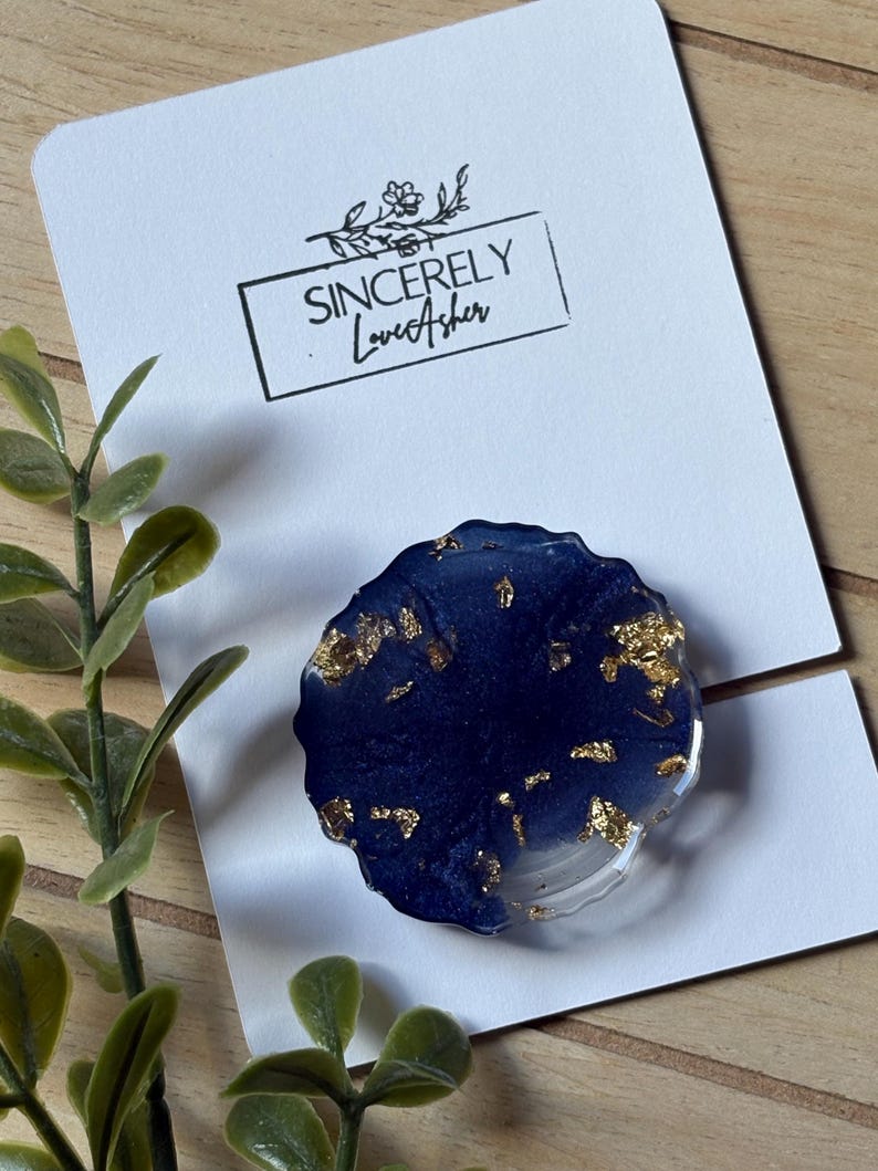 Puede incluir: Un soporte para tel&eacute;fono de resina azul marino con detalles de pan de oro. El agarre redondo tiene una base transparente y est&aacute; sobre una tarjeta blanca con el texto "Sincerely LoveAsher".