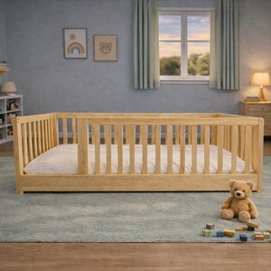 May include: A light-colored wooden toddler bed with a white mattress sits on a light blue rug in a child's bedroom. A stuffed teddy bear and wooden blocks are on the rug. The room has light blue walls, a window, and a dresser.