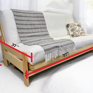 K&ouml;nnte beinhalten: Ein Futon-Sofa aus Holz mit einer wei&szlig;en Matratze und einer grau-wei&szlig; gestreiften Decke. Das Sofa ist 196 cm lang, 94 cm breit und 97 cm hoch. Ein dekoratives Kissen mit floralem Design liegt auf dem Sofa.