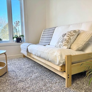 May include: A light wood futon sofa with a white mattress and decorative cushions. A grey and white striped throw is draped over the back. The sofa is in a room with a large window, a round coffee table, and a potted plant.