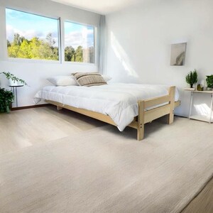 K&ouml;nnte beinhalten: Ein hellfarbiger Holzbettenrahmen mit einer wei&szlig;en Bettdecke und Kissen. Das Bett befindet sich in einem hellen Raum mit einem gro&szlig;en Fenster mit Blick auf eine gr&uuml;ne Landschaft. Ein Teppich in neutraler Farbe bedeckt den Boden.