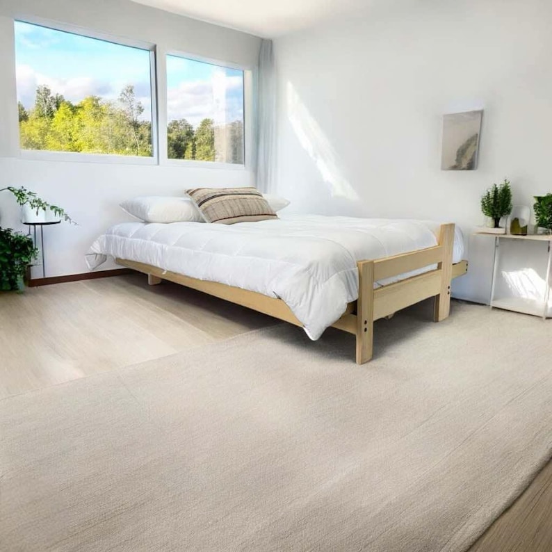 K&ouml;nnte beinhalten: Ein hellfarbiger Holzbettenrahmen mit einer wei&szlig;en Bettdecke und Kissen. Das Bett befindet sich in einem hellen Raum mit einem gro&szlig;en Fenster mit Blick auf eine gr&uuml;ne Landschaft. Ein Teppich in neutraler Farbe bedeckt den Boden.