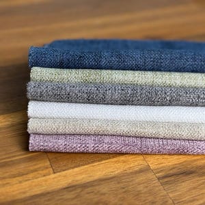 May include: A stack of fabric swatches in various colors, including blue, green, gray, white, beige, and lavender. The fabrics have a textured appearance and are neatly arranged. The swatches are displayed on a wooden surface.