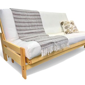 K&ouml;nnte beinhalten: Ein helles Holz-Futon mit wei&szlig;er Matratze und R&uuml;ckenkissen. Eine grau-wei&szlig; gestreifte Decke mit Fransen ist &uuml;ber der R&uuml;ckenlehne drapiert. Ein dekoratives Kissen mit floralem Design liegt auf dem Futon.