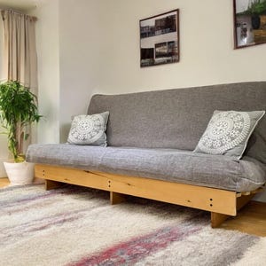 May include: A grey futon sofa with two decorative cushions with a white circular pattern. The sofa is on a light wood frame. A small wooden side table holds a potted plant. A rug with red and purple stripes is on the floor.