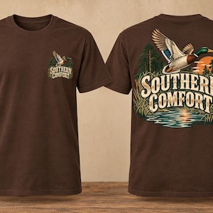May include: A brown short-sleeve t-shirt featuring a small graphic on the front left chest and a large graphic on the back. Both graphics depict a flying mallard duck over a lake with the text Southern Comfort in a stylized beige font.
