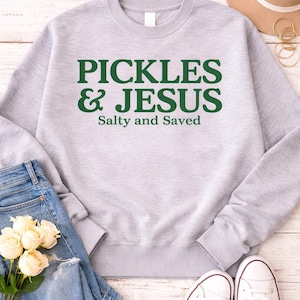 Può includere: Una felpa grigio chiaro con la scritta verde "PICKLES & JESUS" e "Salty and Saved" sotto. L'immagine include jeans blu, scarpe da ginnastica bianche e un mazzo di rose bianche.