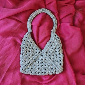 May include: A silver-colored crochet handbag featuring an open-work diamond pattern and a single integrated handle. The bag has a structured, triangular silhouette with a wide base and a narrow top opening, crafted from metallic yarn.