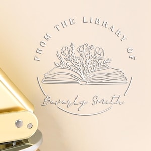 May include: A gold-coloured book embosser with a handle and a circular die. The die is pressed onto a cream-coloured page with the text "FROM THE LIBRARY OF Beverly Smith" surrounding a floral design.