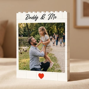 Puede incluir: Un marco de fotos blanco con forma de ladrillo con una foto de un padre y su hijo. El marco tiene las palabras "Daddy & Me" en escritura negra y un corazón rojo. La foto muestra a un hombre sosteniendo a una niña.